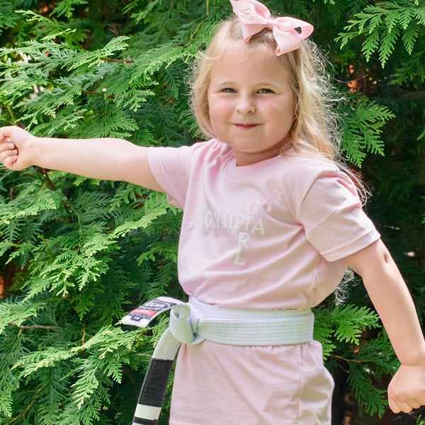 Child in pink shirt and pink shorts with a belt, standing in front of green trees