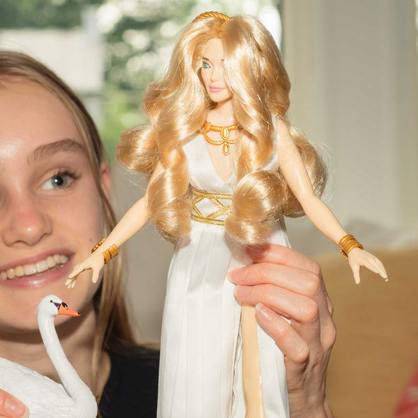 Person holding a doll with long blonde hair and a white dress, with a swan in the foreground.