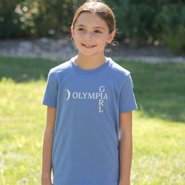 Young girl wearing a blue t-shirt with 'Olympia Girl' text, standing outdoors on grass.