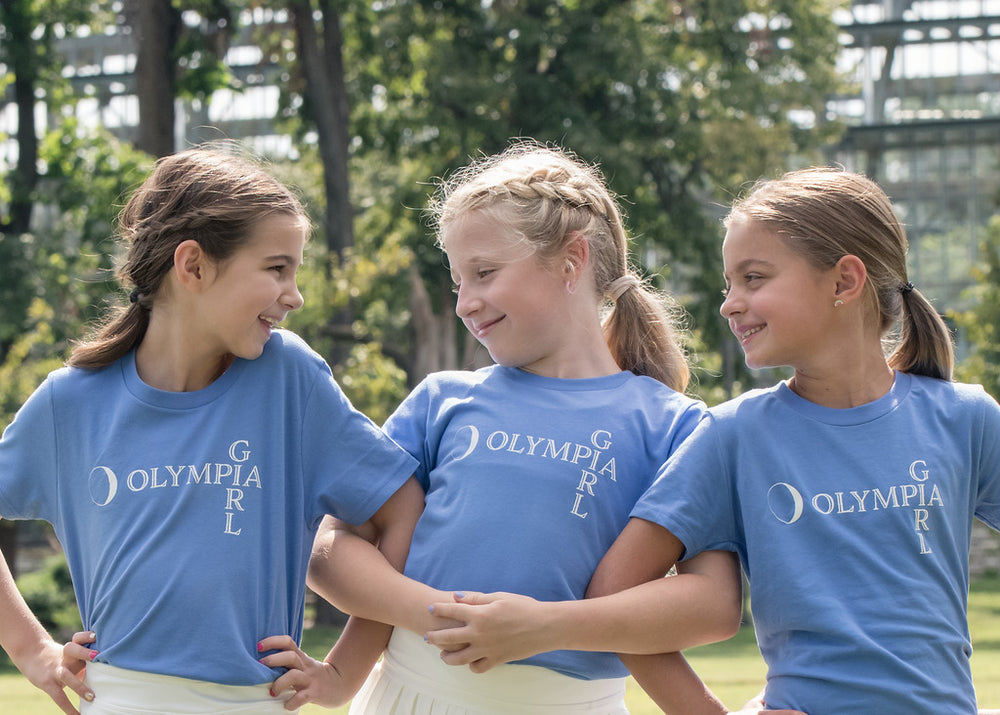 Three girls wearing blue t-shirts with 'Olympia Girl' text, standing outdoors.