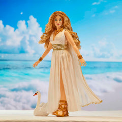 A fashion doll with long wavy blonde hair stands on a beach-like backdrop wearing a flowing white dress with gold trim and matching gold accessories. A white swan figurine is at her side.