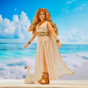 A fashion doll with long wavy blonde hair stands on a beach-like backdrop wearing a flowing white dress with gold trim and matching gold accessories. A white swan figurine is at her side.